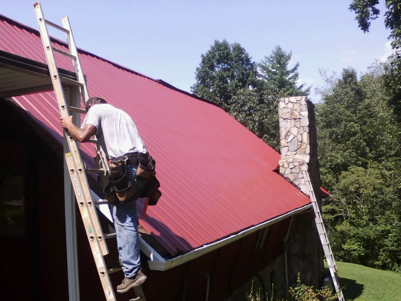 Roofer on a ladder beside a red metal roof and stone chimney — Soffit & Fascia Repair by Roofing Brothers in Hortonville